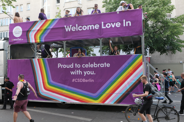 Gruppe von Menschen auf einem geschmückten Doppeldeckerbus bei einer Parade, mit "CSD Berlin"-Banner, umgeben von Bäumen und Gebäuden, Treppe mit Geländer führt zum Bus, einige tragen Mützen, einer hält ein Fahrrad.