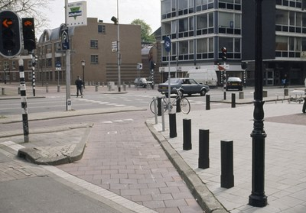 Eine Stadtstraße mit einem geparkten Fahrrad, fahrenden Fahrzeugen, Fußgängern auf dem Gehweg, Versorgungsmasten, Verkehrsampeln, Straßenschildern, Bäumen, Gebäuden mit Fenstern und einem klaren blauen Himmel.