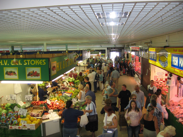 Ein belebter Marktplatz mit Menschen, die Obst und Gemüse anschauen und kaufen, unter Deckenbeleuchtung.