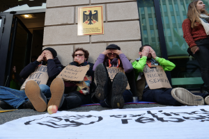 Eine Gruppe von Menschen sitzt vor einem Gebäude auf dem Boden und hält Schilder, mit einem Banner und einem Apfel auf der Straße, und eine Frau steht rechts daneben und sieht besorgt aus.