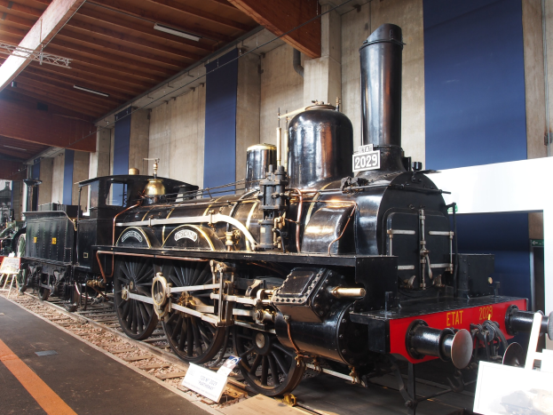 Eine große, alte Dampflokomotive in einem Museums Setting umgeben von Eisenbahnschienen, Informationsschildern, Ständen und anderen Ausstellungen, mit einer Wand- und Deckenbeleuchtung im Hintergrund.