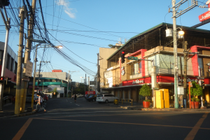 Eine Stadtstraße mit Autos, Fußgängern, Versorgungsmasten, Ampeln, Gebäuden mit Schildern, Topfpflanzen und einer bewölkten Himmel.