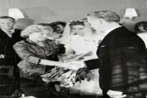 Ein Schwarz-Weiß-Foto von einer Braut und einem Bräutigam, die sich die Eheringe reichen, wobei die Braut ein weißes Kleid und einen Schleier trägt und einen Blumenstrauß hält, und der Bräutigam einen Anzug und eine Krawatte trägt, mit Wandlampen im Hintergrund.