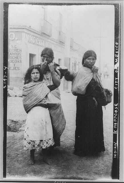 Drei Frauen, die nebeneinander auf einer Straße in Mexiko-Stadt stehen, mit Gebäuden im Hintergrund, auf einem Schwarz-Weiß-Foto.