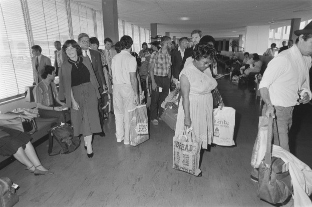 Ein Schwarz-Weiß-Bild von Reisenden in einem Flughafen, einige sitzen auf Bänken, einige stehen, mit einem Mann und einer Frau, die Koffer halten und Fensterläden auf der linken Seite.