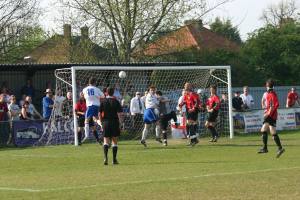 Spieler sind in ein Fußballspiel auf einem Feld mit einem Tor involviert, während Zuschauer dahinter stehen, mit Bäumen und Häusern im Hintergrund.