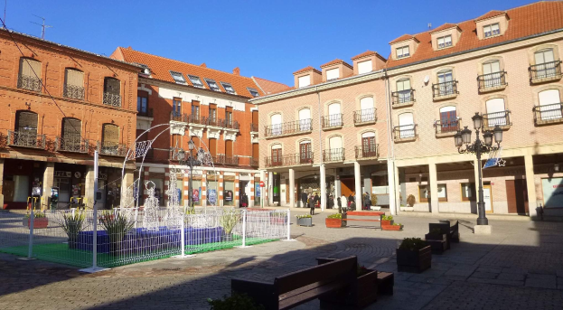 Stadtplatz mit einem zentralen Brunnen, umgeben von Bänken, Topfpflanzen, einem Metallzaun, Straßenlaternen, Menschen, Schildern, Gebäuden mit Fenstern und einem bewölkten Himmel.