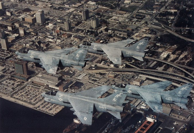 Vier Kampfflugzeuge fliegen über einer Stadt mit sichtbaren Gebäuden, Straßen, Fahrzeugen und Wasser, mit Text am unteren Bildrand.