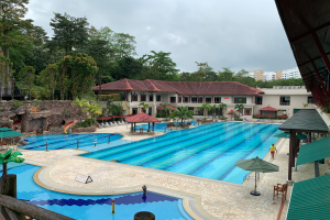 Ein großes Schwimmbecken im Ritz-Carlton, Kota Kinabalu, voller Menschen, umgeben von Schirmen, Stühlen, Tischen, Pflanzen, Bäumen, Felsen und Gebäuden, mit einem Schuppen auf der rechten Seite und dem Himmel im Hintergrund.