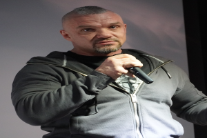 Ein Mann in einer schwarzen Kapuzenjacke, einem schwarzen Anzug und einem weißen Hemd, mit zurückgekämmtem Haar, hält ein Mikrofon und steht selbstbewusst vor einer Wand.