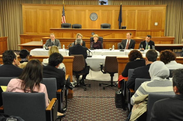 Ein Versammlungsraum mit auf Stühlen sitzenden Menschen, zwei ausländischen Flaggen, einem orangen Tisch, Wasserflaschen und einem Mikrofon.