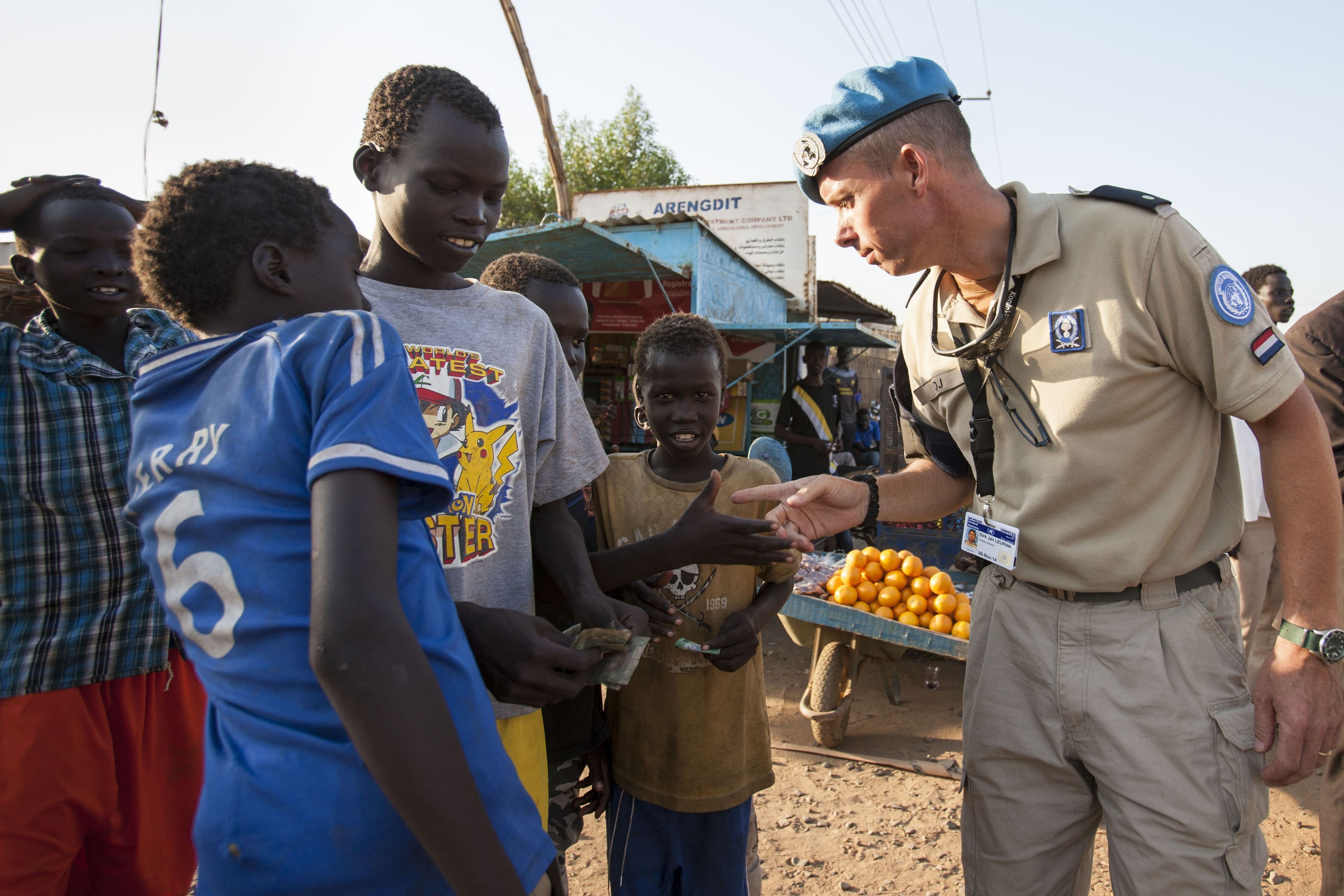 Polizist in Uniform spricht zu Kindern neben einem Lastwagen mit Orangen, mit Scheunen, Bäumen und einem klaren Himmel im Hintergrund.