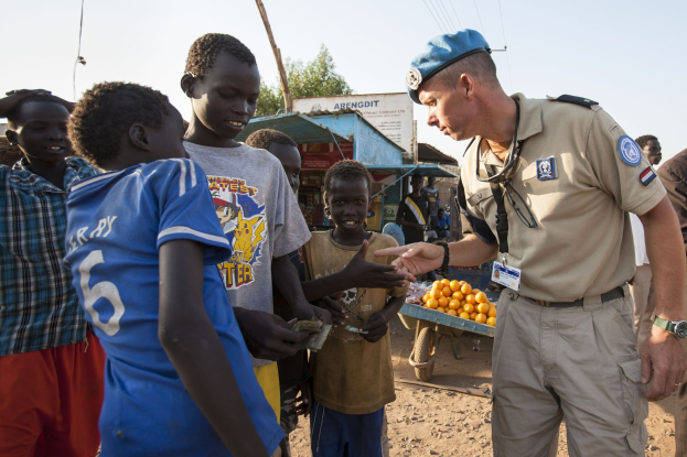 Polizist in Uniform spricht zu Kindern neben einem Lastwagen mit Orangen, mit Scheunen, Bäumen und einem klaren Himmel im Hintergrund.