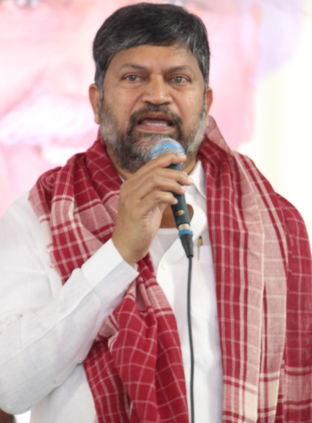 A man in a white shirt and red shawl speaks into a microphone at a press conference, with a banner featuring a person's image in the background.