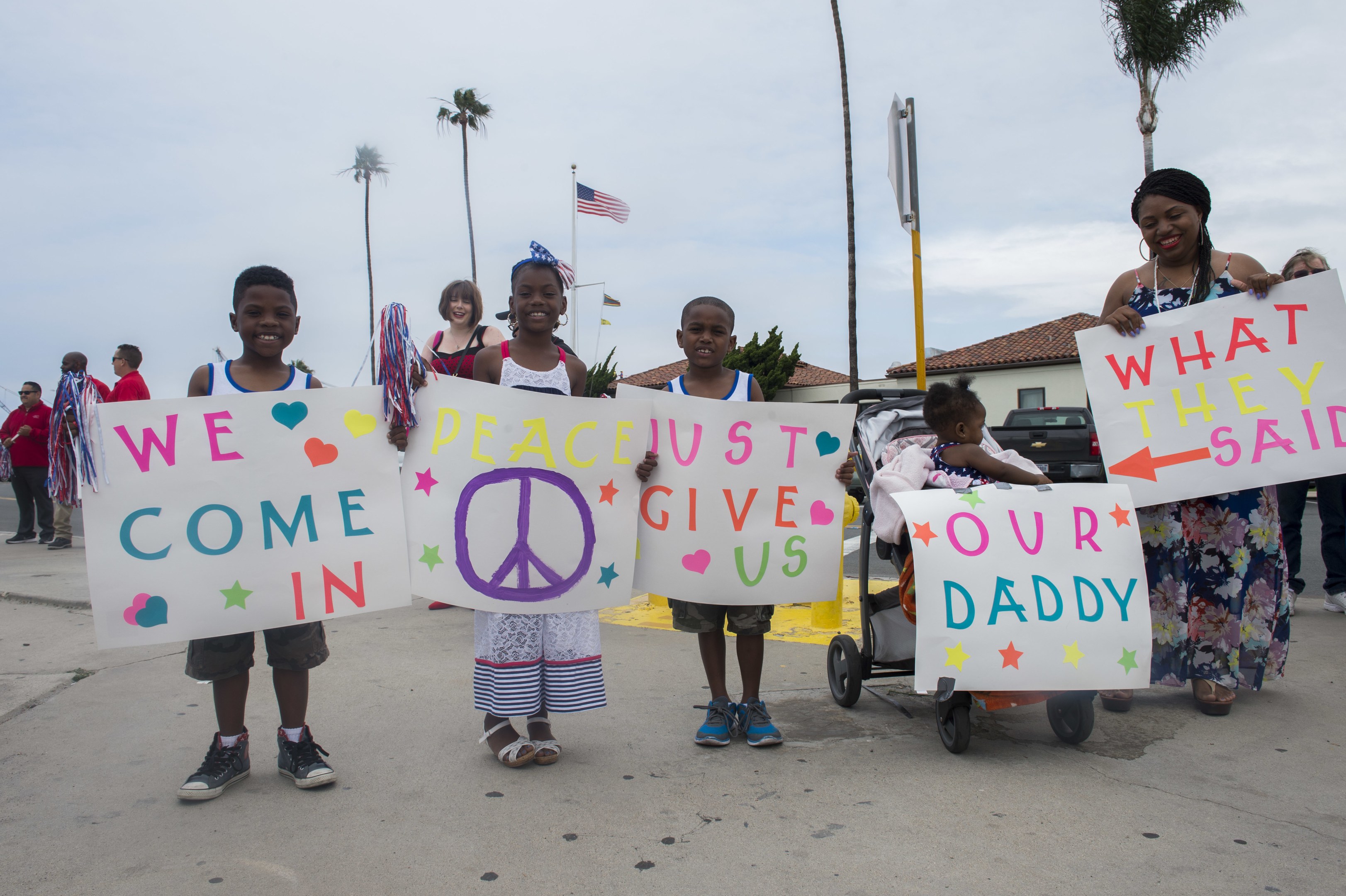 Eine Gruppe von Kindern hält Schilder in der Hand und steht vor einem Gebäude, während eine Frau einen Kinderwagen schiebt, in dem ein Baby sitzt; auf einem Schild steht "Wir kommen in Frieden, gebt uns unseren Daddy."