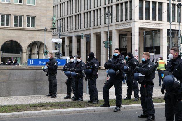 Eine Gruppe von Polizisten in schwarzen Uniformen und Masken steht vor einem Gebäude mit Glasfenstern und Säulen in Berlin, Deutschland, einige halten Helme, mit Laternen, Verkehrszeichen, Texttafeln, einer Statue, anderen Menschen und grasbewachsenem Boden im Hintergrund.