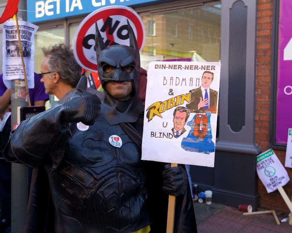 A man in a Batman costume holds a placard in the foreground, with two more placards leaning against a wall, scattered objects on the floor, and people, a flag, a pole, a poster, a banner, and a glass wall visible in the background.