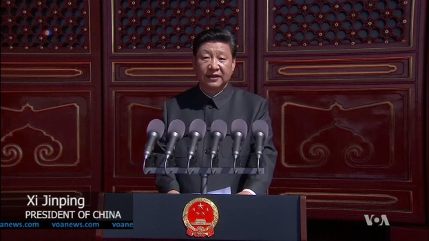 Xi Jinping steht an einem Podium mit Mikrofonen und einem Logo, während er eine Rede in der Großen Halle des Volkes in Peking hält, mit einer hölzernen Wand im Hintergrund.
