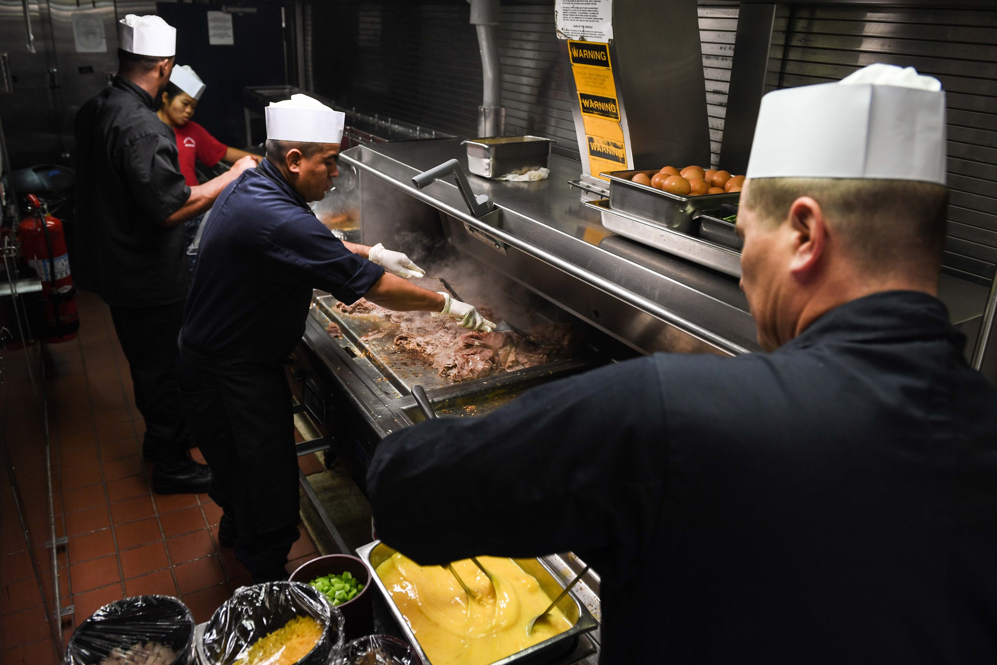 Four chefs in black uniforms and white caps grilling food at a table with trays of items, with a fire extinguisher visible in the background.