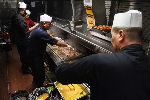 Four chefs in black uniforms and white caps grilling food at a table with trays of items, with a fire extinguisher visible in the background.