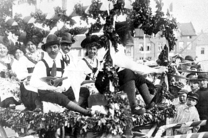 Eine Gruppe von Menschen in traditioneller bayrischer Tracht fährt auf einem mit Laub geschmückten Festwagen mit, einige halten Musikinstrumente, im Hintergrund sind Gebäude und ein klarer Himmel zu sehen, dargestellt in Schwarz-Weiß.