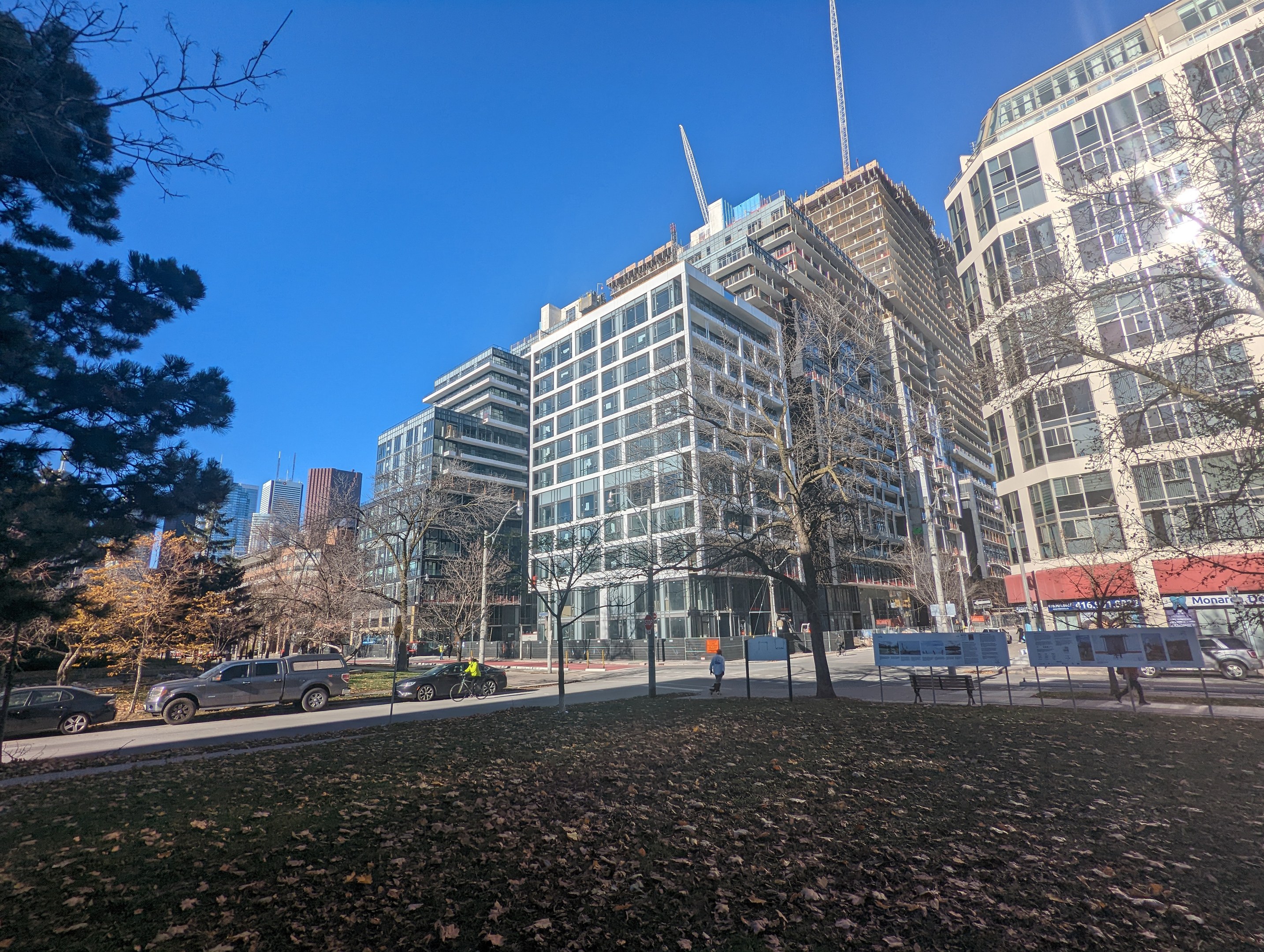 A city street in Toronto with tall buildings, moving vehicles, pedestrians on the sidewalk, trees, and scattered dry leaves, under a visible sky.