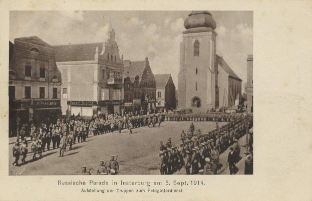 Schwarzes Foto einer Parade in Insterburg 1914 mit Menschen, die gehen und stehen, Gebäuden im Hintergrund und Wolken am Himmel.