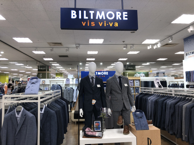 Innenansicht eines Biltmore Vis Viva Bekleidungsgeschäfts mit Schaufensterpuppen in Anzügen, aufgehängten Kleidungsstücken, einem Tisch mit einer Blumenvase, einer deckenmontierten Texttafel und einer Beleuchtung von oben.