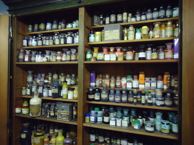 Ein hölzerner Schrank voller ordentlich arrangierter Medikamentenflaschen, mit einer Tür auf der linken Seite.