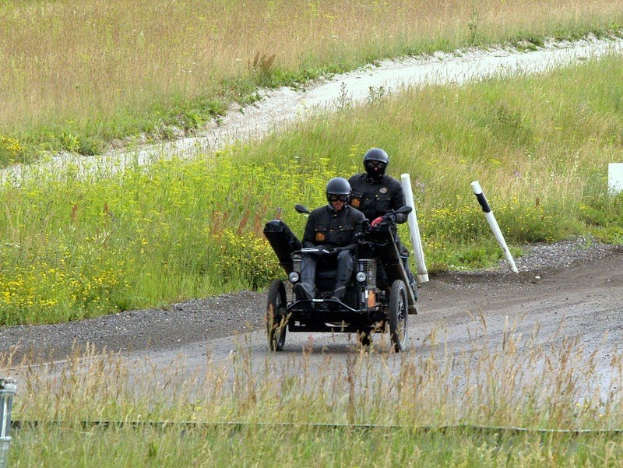 Zwei Polizisten in schwarzen Uniformen und Helmen fahren auf einem dreirädrigen Motorrad über einen mit Gras und Pflanzen gesäumten Schotterweg, im Hintergrund sind weiße Pfosten zu sehen.