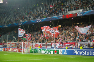 Ein Fußballspiel wird in einem gut beleuchteten Stadion mit einer großen Zuschauermenge, grünem Rasen, einem Torpfosten mit Bannern und einem Bildschirm in der oberen linken Ecke gespielt.