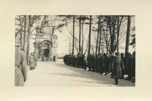 Ein Schwarz-Weiß-Foto einer Gruppe von Menschen, die vor einem Gebäude mit einem Torbogen und Bäumen an der Straße stehen, mit einem Text unten, der lautet: "Die Trauerfeier für die Opfer der Berliner Mauer."