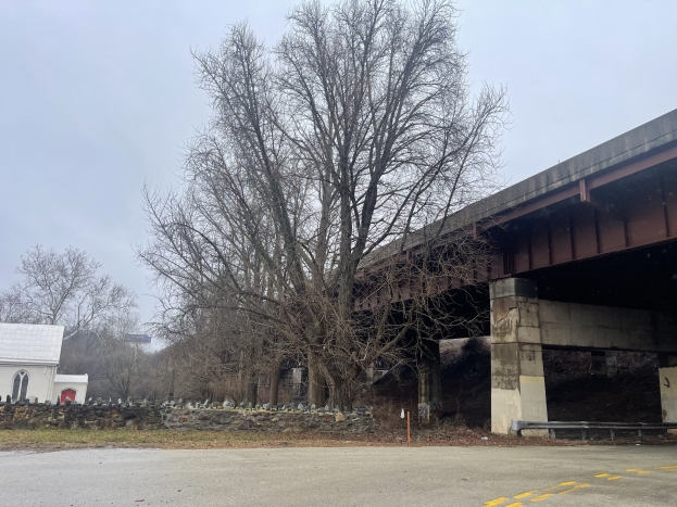Eine Brücke in Reparatur über einer Straße, mit einem Baum in der Mitte umgeben von Steinen, Gras, einem Haus, einem Schild, einem Straßenpfahl, einer Baumgruppe und einem bewölkten Himmel.