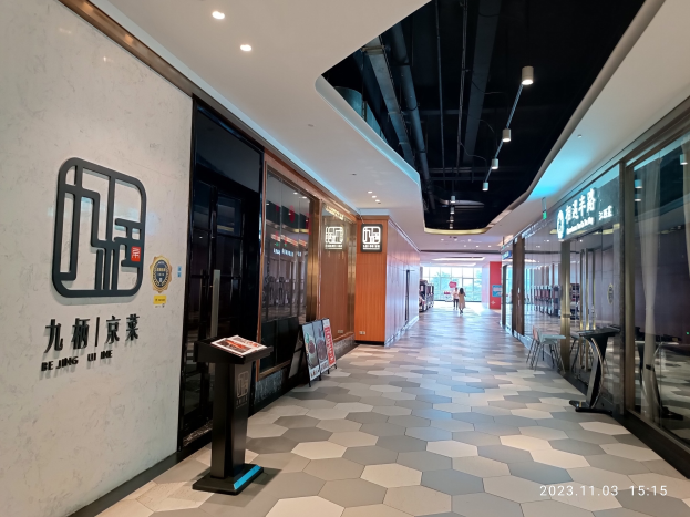 Lobby des Beijing U Me Einkaufszentrums mit einem großen beleuchteten Schild, Namen und Informationsschildern, Stühlen und Menschen, die gehen.