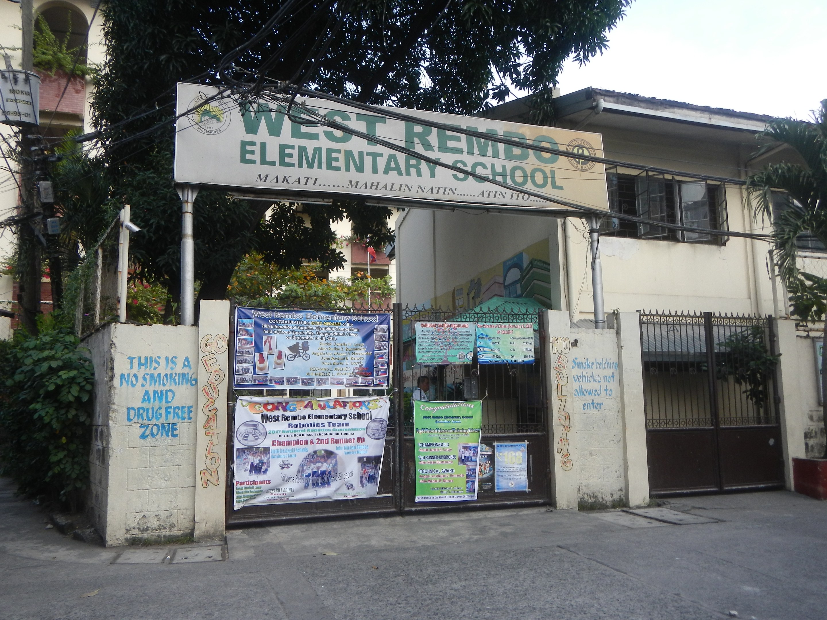 Außenansicht der West Rembo-Grundschule in Manila, Philippinen, mit einem Gebäude mit Fenstern, einem Tor, einer Tafel mit Text, Plakaten an der Wand, Pflanzen, Bäumen, einem Straßenpfosten, Drähten und einem bewölkten Himmel.