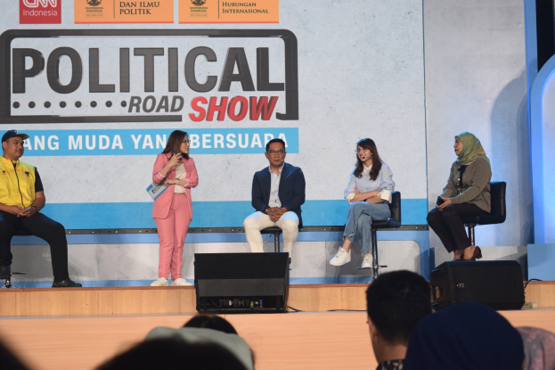 Eine Frau mit einem Mikrofon steht in der Mitte einer Bühne, während das Publikum darunter zusieht, mit einem 'Political Road Show 2019'-Plakat im Hintergrund.