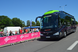 Ein schwarzer und grüner Bus fährt auf einer Straße neben einer Menge Menschen, einige tragen Möppen, mit einem Banner auf der linken Seite, Bäumen und einem klaren blauen Himmel im Hintergrund.
