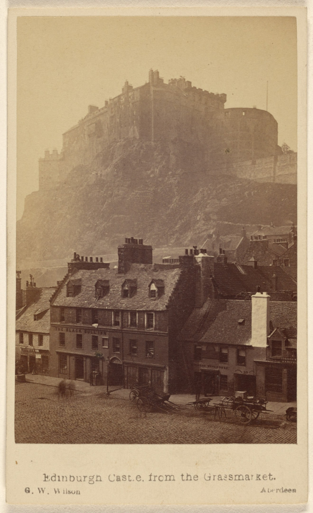 Altes Foto von Edinburgh Castle aus der Grassmarket-Sicht, mit Gebäuden und Karren im Vordergrund und einem Hügel im Hintergrund, mit Text unten.
