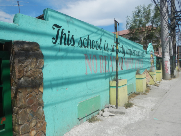 Grüne Wand mit Graffiti-Schriftzug "Dies ist keine Stadt" umgeben von Gebäuden, Bäumen, Strommasten, Drähten und anderen städtischen Elementen unter einem bewölkten Himmel.