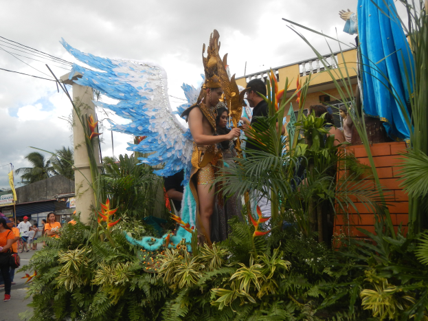 Eine Gruppe von Menschen geht eine Straße entlang neben einem schwimmenden Aufbau, der mit Pflanzen und Blumen geschmückt ist, mit kostümierten Personen auf dem Aufbau, Gebäuden, Bäumen, Strommasten mit Drähten und einem bewölkten Himmel im Hintergrund.