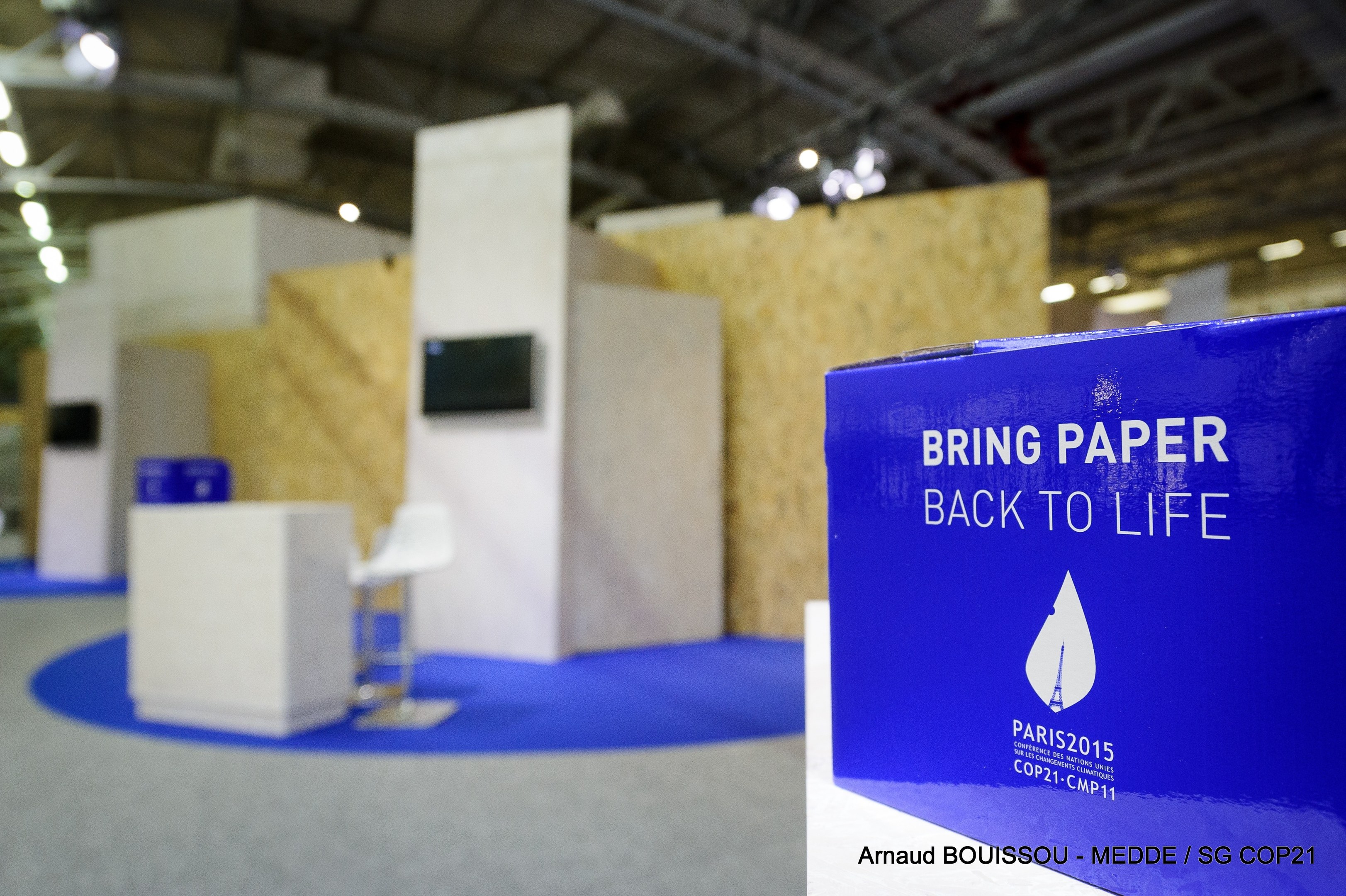 Messestände mit einem blauen und weißen 'Bring Paper Back to Life'-Schild auf der rechten Seite, mit unscharfen Stühlen, Tischen, einem wandmontierten Fernseher und Deckenleuchten im Hintergrund.