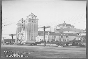 Ein Schwarz-Weiß-Foto einer römisch-katholischen Kathedrale und eines Bischofssitzes mit Gebäuden, Bäumen, Polen, Drähten, Menschen und einer Straße im Vordergrund und dem Himmel im Hintergrund, mit Text am unteren Rand des Bildes.