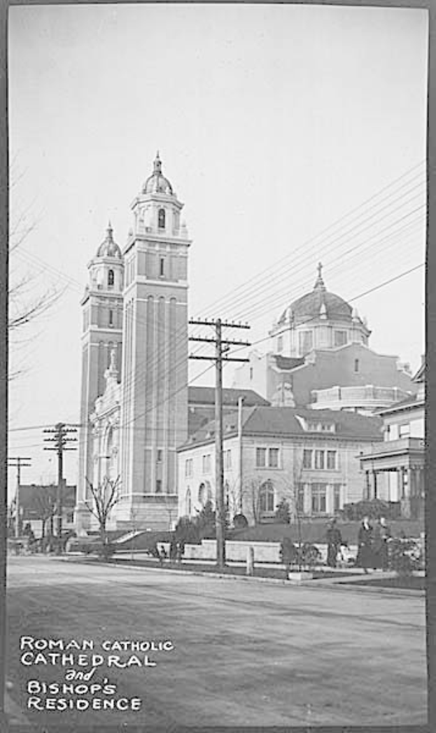 Ein Schwarz-Weiß-Foto einer römisch-katholischen Kathedrale und eines Bischofssitzes mit Gebäuden, Bäumen, Polen, Drähten, Menschen und einer Straße im Vordergrund und dem Himmel im Hintergrund, mit Text am unteren Rand des Bildes.