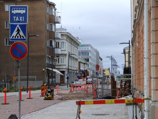 Stadtstraße mit Gebäuden, Straßenmästchen, Lichtern, Schildern, Absperrgittern, Fahrzeugen, Barrieren, Bäumen und einem bewölkten Himmel.