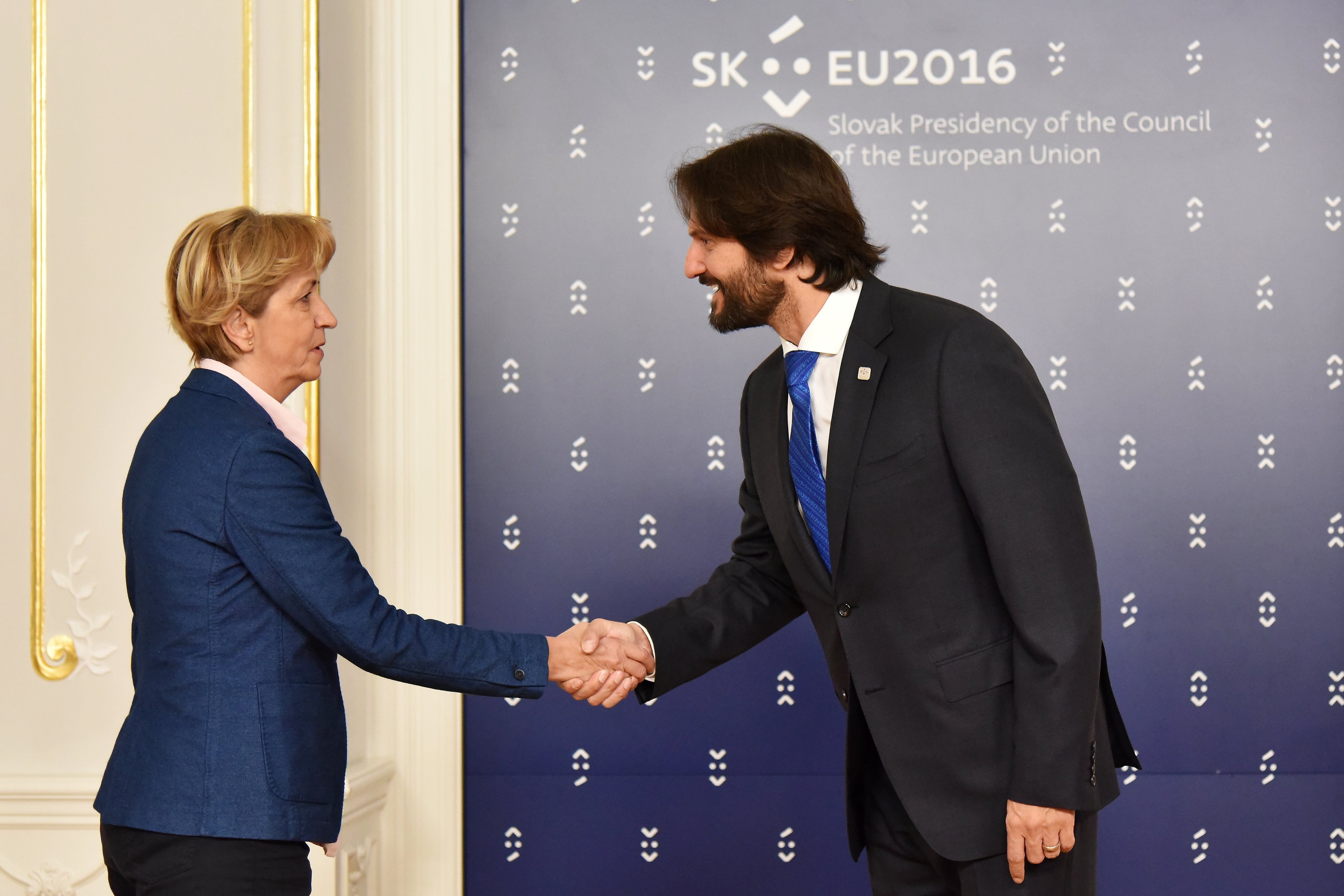 Bundeskanzlerin Angela Merkel und Bundeskanzler Joachim Gauck schütteln sich vor einer Wand mit einem 'EU-Gipfel'-Banner und einer Tür im Hintergrund die Hände, beide lächeln.
