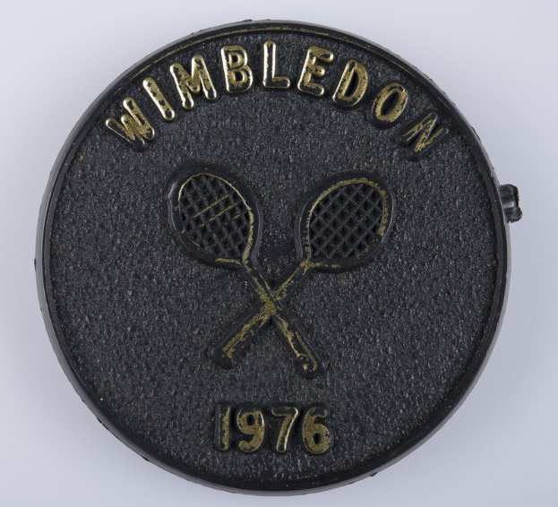 Schwarze Münze mit "Wimbledon 1976" und zwei gekreuzten Tennisrackets auf einem weißen Hintergrund.