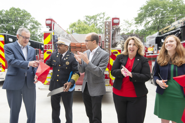 Gruppe von Menschen bei einer Eröffnungszeremonie für die Alexandria Fire Department, die vor einem Feuerwehrauto klatschen und lächeln, vor einem Hintergrund mit Bäumen und einem blauen Himmel.