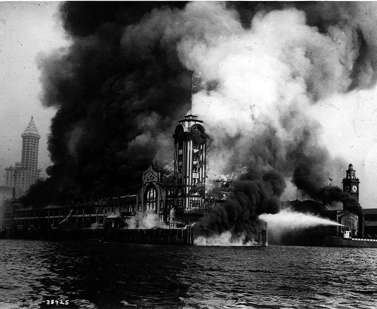 Schwarzes und weißes Foto eines großen brennenden Gebäudes mit Rauch, der daraus aufsteigt, einem Gewässer im Vordergrund und anderen Gebäuden im Hintergrund.