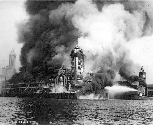 Schwarzes und weißes Foto eines großen brennenden Gebäudes mit Rauch, der daraus aufsteigt, einem Gewässer im Vordergrund und anderen Gebäuden im Hintergrund.