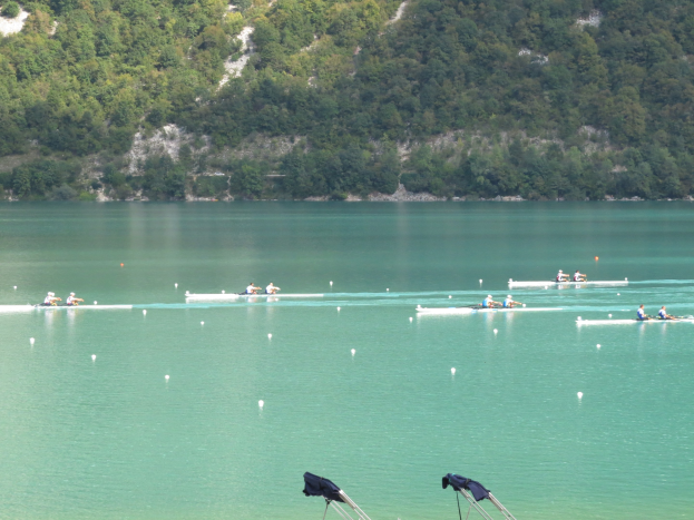 Eine Gruppe von Menschen, die bei einem Wettbewerb auf einem Gewässer mit Bäumen und einem Hügel im Hintergrund rudern, während Kleidung auf Ständern darunter hängt.